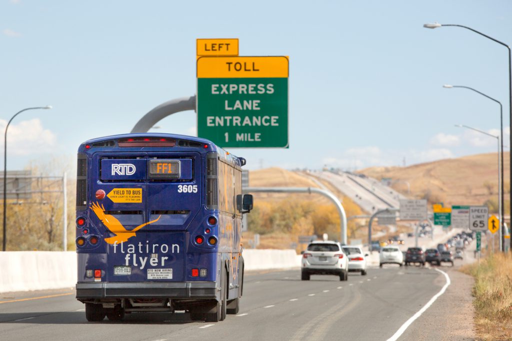 Shot of the FlatIron Flyer bus from behind as the bus gets onto the highway. Toll lane signage is right above the bus.
